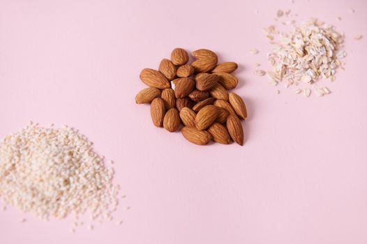 Selective focus on a handful of raw organic almond nuts, near scattered sesame seeds and oat-flakes on a pink background, copy advertising space. View from above. Flat lay. Still life. Superfood.