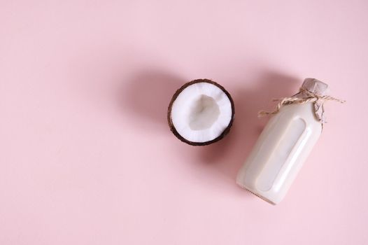 Flat lay composition with a half of raw coconut, lying next to a glass bottle with organic vegan plant based milk, isolated on pink background with copy space for advertising text. Still life
