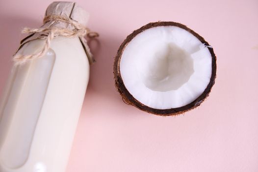 Close-up of a glass bottle of organic raw vegan milk with a craft paper tying with a bow on a lid, and a half of coconut, isolated over pink background with copy ad space. Healthy food and drink