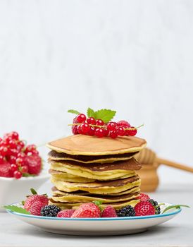 Stack of baked pancakes with fruits in a round plate on a white table