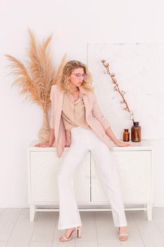 a stylish slender woman in glasses, of 30-40 years old with blond hair stands in a light pink jacket and white trousers in the interior of a white room, near a chest of drawers, looks to the side. Copy space. Vertical