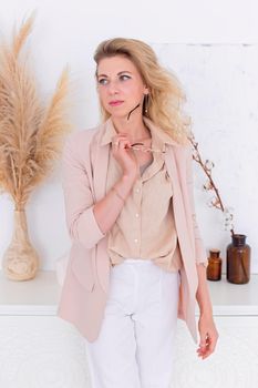 A beautiful woman with blond hair, stands in a light pink jacket in the interior of a white room, look to the side . Copy space. Vertical