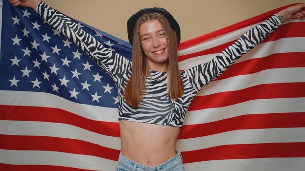 Lovely young woman in crop top waving and wrapping in American USA flag, celebrating, human rights and freedoms. Independence day. Adult stylish girl isolated alone on beige studio background indoors