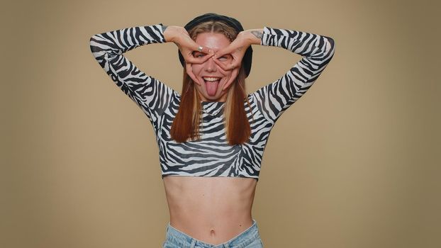 Lovely pretty funny woman in crop top making playful silly facial expressions and grimacing, fooling around, showing tongue. Adult stylish female girl isolated alone on beige studio background indoors