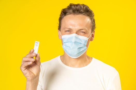 Happy young man showing test in hand wearing medical face mask celebrating victory pandemic coronavirus or monkeypox. Man in white t-shirt and medical mask on yellow background.