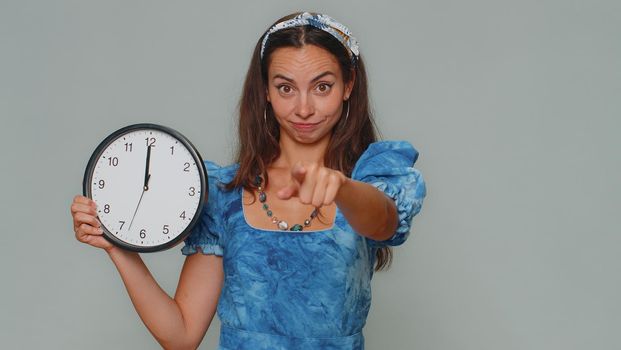 It is your time. Pretty young woman in blue princess dress showing time on wall office clock, ok, thumb up, approve, pointing finger at camera. Stylish female girl on gray studio background indoor