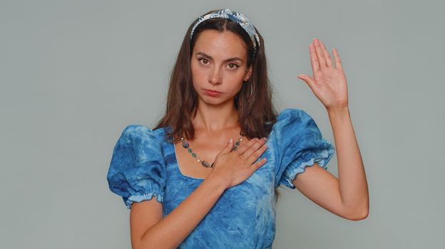 I swear to be honest. Sincere responsible pretty young woman raising hand to take oath, promising to be honest and to tell truth, keeping hand on chest. Adult stylish girl isolated on gray background