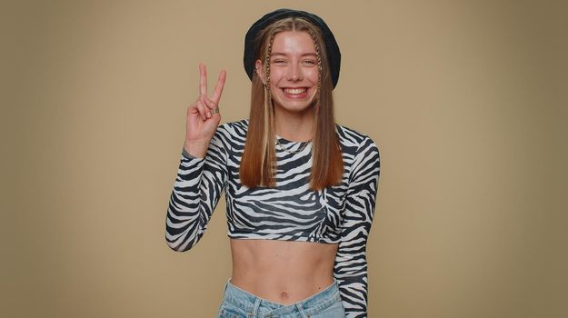 Lovely happy young woman showing victory sign, hoping for success and win, doing peace gesture, smiling with kind optimistic expression. Adult stylish girl isolated on beige studio background indoors