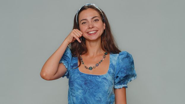 Call me, here is contact number. Lovely pretty young woman looking at camera doing phone gesture like says hey you call me back. Adult stylish female girl isolated on gray studio background indoors