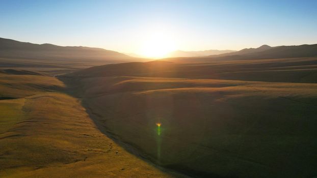 Bright yellow dawn with a red tint in the mountains. The green hills with grass turned red. A small stream flows through the gorge, horses graze. Clear blue sky. View from top. Assy Plateau Kazakhstan