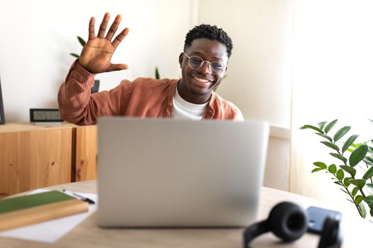 Black young man in video call taking online classes. Greeting classmates. Waving hand towards laptop. E-learning concept. Working from home. Video call concept.