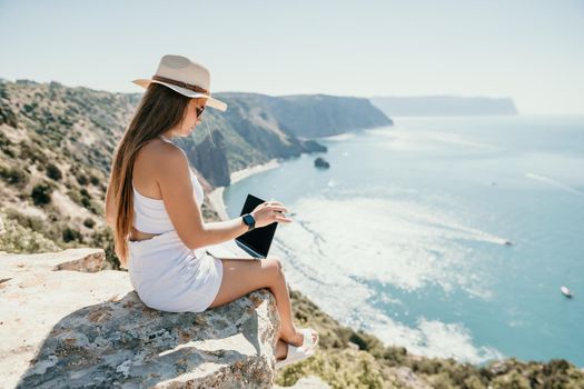 Successful business woman in yellow hat working on laptop by the sea. Pretty lady typing on computer at summer day outdoors. Freelance, travel and holidays concept.
