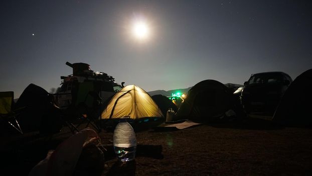 A big bright moon in the starry sky. There is a tent camp. The yellow tent is glowing. Several cars with things. Transparent plastic bottle and bags on the ground. Lanterns are lit. Assy, Kazakhstan
