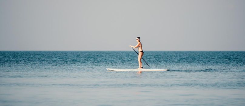 Silhouette of woman standing, surfing on SUP board, confident paddling through water surface. Idyllic sunset or sunrise. Sports active lifestyle at sea or river.