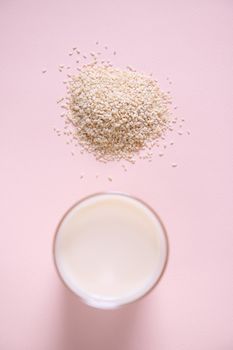 Top view. Selective focus on a heap of sesame seeds, scattered on pink background, near drinking glass of healthy nutritious white non dairy drink. Alternative wholesome organic plant based milk from