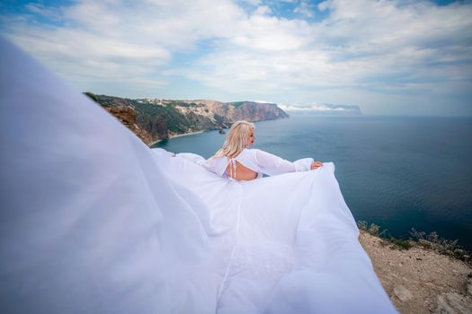 Blonde with long hair on a sunny seashore in a white flowing dress, rear view, silk fabric waving in the wind. Against the backdrop of the blue sky and mountains on the seashore