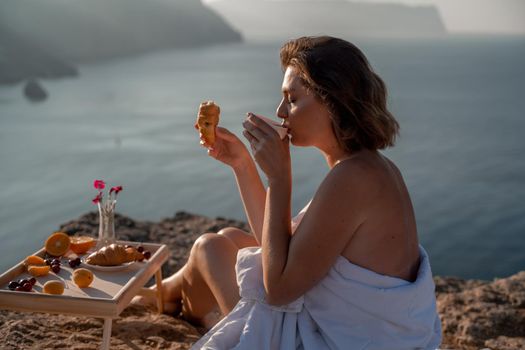 Woman covered with a blanket of bed relaxing and watching the seascape at sunrise. She holds a cup of coffee in her hand in front of her is a table with fruits and croissants. Wanderlust and freedom concept scene