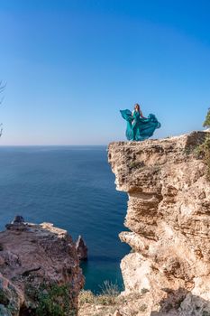 A girl with loose hair in a long mint dress descends the stairs between the yellow rocks overlooking the sea. A rock can be seen in the sea. Sunny path on the sea from the rising sun.