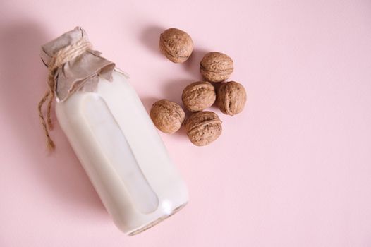 Still life with a plant based milk in a glass bottle, alternative non dairy drink and a heap of organic wholesome walnuts on a pink background with advertising copy space. Healthy food and drink