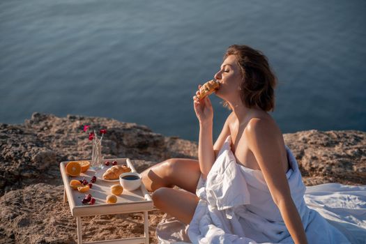 Woman covered with a blanket of bed relaxing and watching the seascape at sunrise. She holds a cup of coffee in her hand in front of her is a table with fruits and croissants. Wanderlust and freedom concept scene