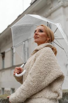 The blonde stands under a transparent umbrella during the rain. The fall season. Rear view. The woman is dressed in a black lace dress, her hair pulled back in a ponytail