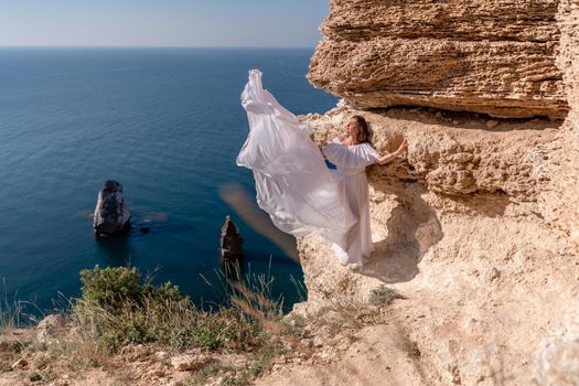 A beautiful young woman in a white light dress with long legs stands on the edge of a cliff above the sea waving a white long dress, against the background of the blue sky and the sea