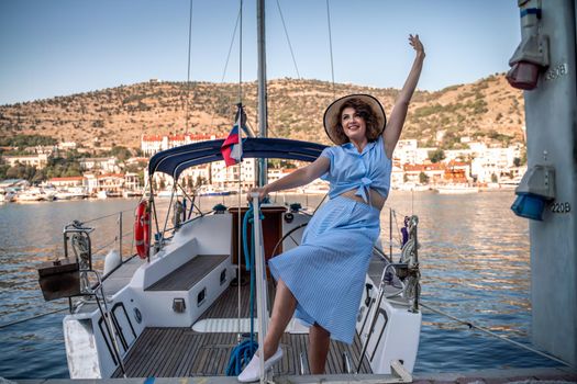 A young happy woman in a blue dress and hat stands near the seaport with luxury yachts. Travel and vacation concept.