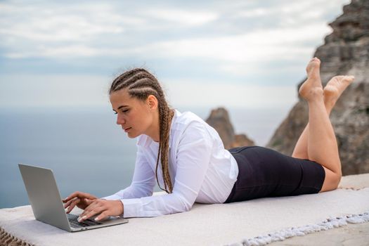 A woman is lying and typing on a laptop keyboard on a terrace with a beautiful sea view. Wearing a white blouse and black skirt. Freelance travel and vacation concept, digital nomad