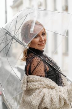 The blonde stands under a transparent umbrella during the rain. The fall season. Rear view. The woman is dressed in a black lace dress, her hair pulled back in a ponytail