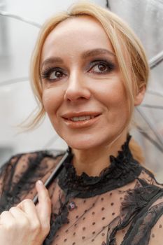 The blonde stands under a transparent umbrella during the rain. The fall season. Rear view. The woman is dressed in a black lace dress, her hair pulled back in a ponytail