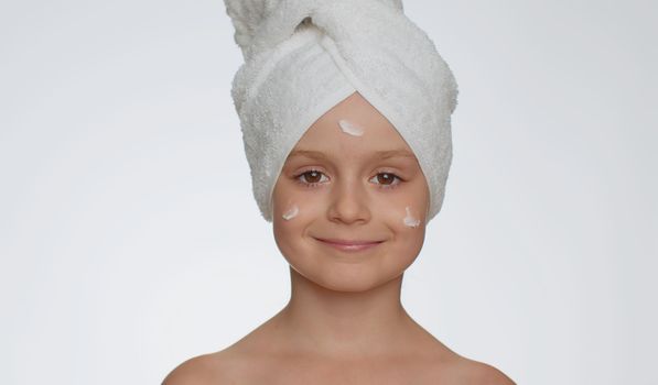 Lovely young child girl after bath in towel on head applying cleansing moisturizing cream. Teenager kid face skincare healthy treatment, natural creme cosmetics. Female portrait. Perfect fresh clean