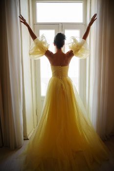 A woman's silhouette in a golden luxurious dress against the background of a window holds a curtain with her hands. Elegant lady in a yellow long silk dress with bare back, back view