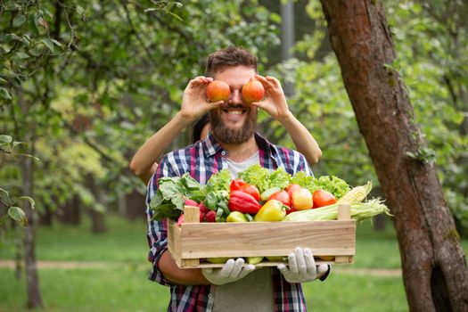 Funny shot of farmer with two apple as glasses and wooden box of vegetables in garden, autumn summer organic harvest concept