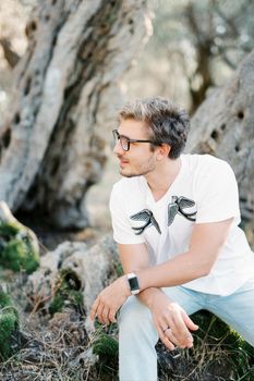 Young man with glasses sits turned sideways near a tree. High quality photo