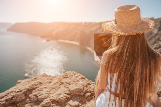 Successful business woman in yellow hat working on laptop by the sea. Pretty lady typing on computer at summer day outdoors. Freelance, travel and holidays concept.