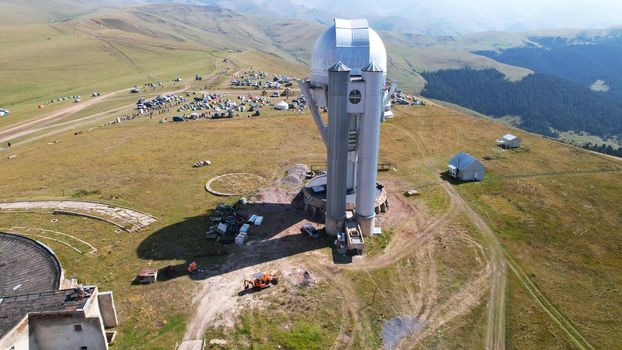 The Assy-Turgen Observatory is high in the mountains. There is a tent camp next to the observatory. Large cumulus clouds in a blue sky. Yellow-green hills, forest in places. Top view from a drone