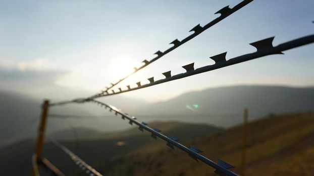 Sharp barbed wire on the fence with a blurred background of mountains. A bright yellow sun in a blue sky. Gray clouds. The high hills are covered with yellow-green grass. Restricted area. Background