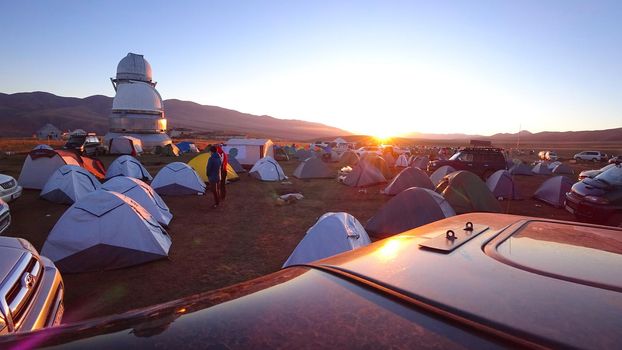 Sunrise over the camp near the Assi Observatory. The sun comes out from behind the high hills. Red-yellow color. Lots of tents and cars. The guy launches the drone. Telescopes are covered with domes