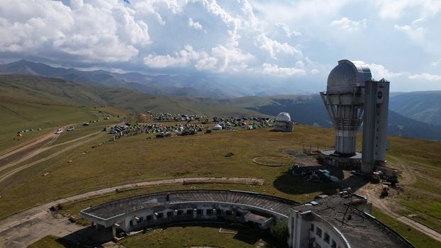 The Assy-Turgen Observatory is high in the mountains. There is a tent camp next to the observatory. Large cumulus clouds in a blue sky. Yellow-green hills, forest in places. Top view from a drone