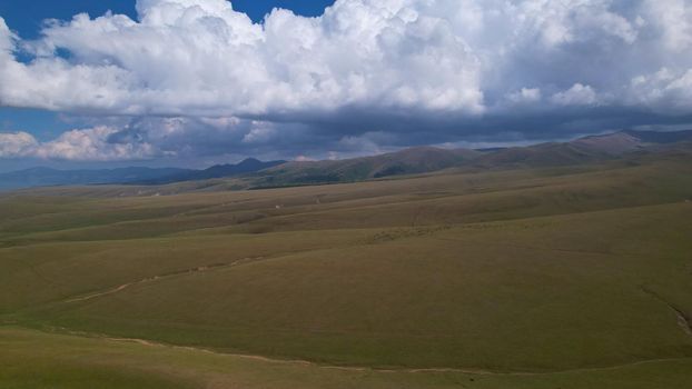 Big white clouds over green hills and mountains. Top view from the drone on endless fields. Roads are visible in places, herds of animals graze. A tent camp has been set up. Coniferous trees in gorge