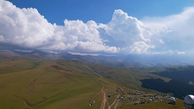 Big white clouds over green hills and mountains. Top view from the drone on endless fields. Roads are visible in places, herds of animals graze. A tent camp has been set up. Coniferous trees in gorge