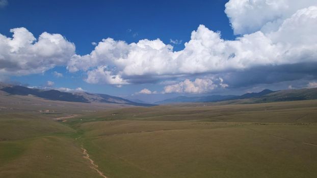 Big white clouds over green hills and mountains. Top view from the drone on endless fields. Roads are visible in places, herds of animals graze. A tent camp has been set up. Coniferous trees in gorge