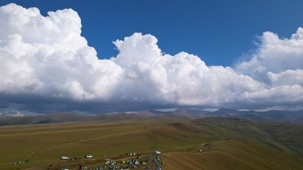 Big white clouds over green hills and mountains. Top view from the drone on endless fields. Roads are visible in places, herds of animals graze. A tent camp has been set up. Coniferous trees in gorge
