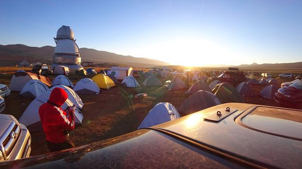 Sunrise over the camp near the Assi Observatory. The sun comes out from behind the high hills. Red-yellow color. Lots of tents and cars. The guy launches the drone. Telescopes are covered with domes