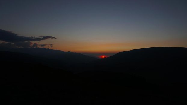 Sunset sun over yellow-green hills. Small clouds are illuminated by the orange-red rays of the outgoing sun. A light haze in the gorge. Coniferous trees grow. Clear sky. Assi-Turgen Plateau Kazakhstan