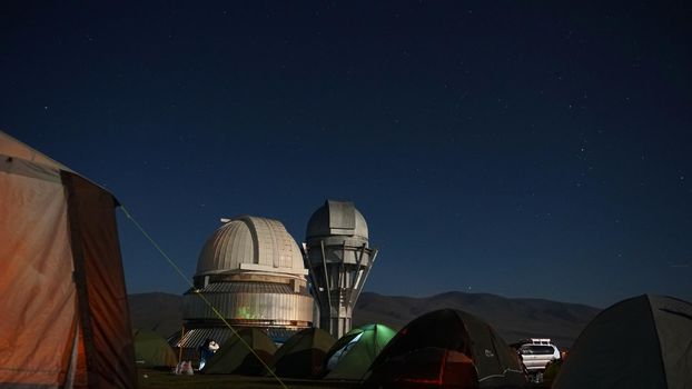 A camp with colored tents near the observatory. The Perseid starfall. A large teleskom watches the starry night sky. Meteorites and planes fly by, leaving traces in the sky. The stars are burning.