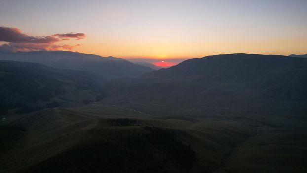 Red rays of the sun at sunset among the mountains. Green grass turns yellow, in places a small forest. Tourists walk in the hills. A river runs in the distance. The clouds are highlighted in pink.