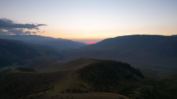 Red rays of the sun at sunset among the mountains. Green grass turns yellow, in places a small forest. Tourists walk in the hills. A river runs in the distance. The clouds are highlighted in pink.