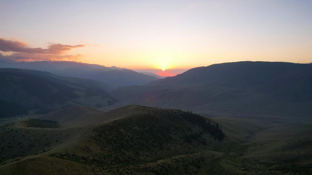 Red rays of the sun at sunset among the mountains. Green grass turns yellow, in places a small forest. Tourists walk in the hills. A river runs in the distance. The clouds are highlighted in pink.