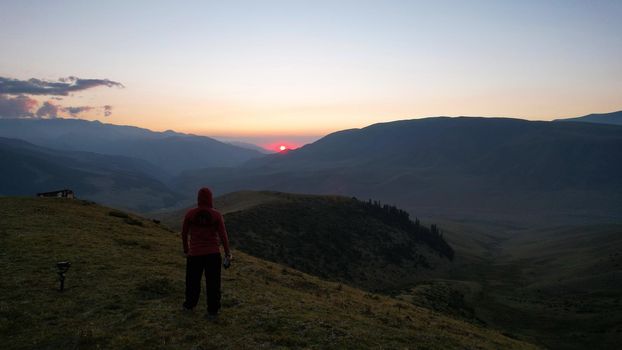 A guy in the mountains is watching the sunset. An epic orange-red sunset is reflected on small clouds and green hills. Bushes and forest grow in places on mountains. The guy is standing with his back